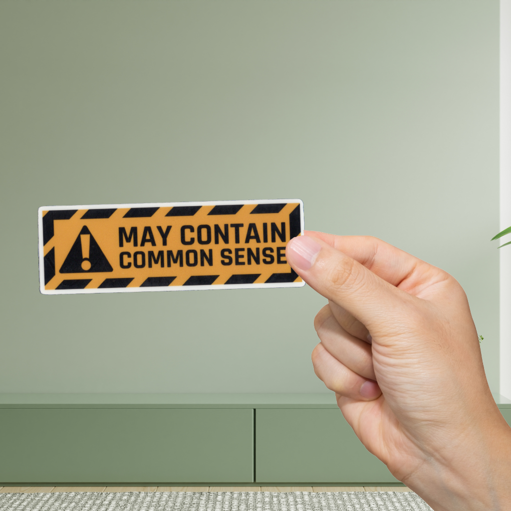 Hand holding a humorous warning sticker with text 'May contain common sense' against a green wall.