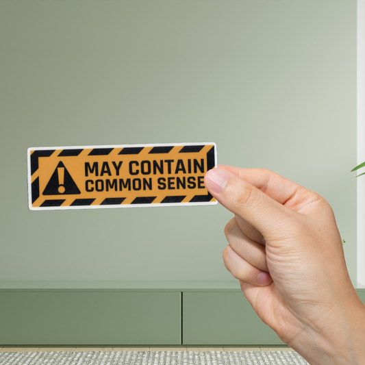 Hand holding a humorous warning sticker with text 'May contain common sense' against a green wall.