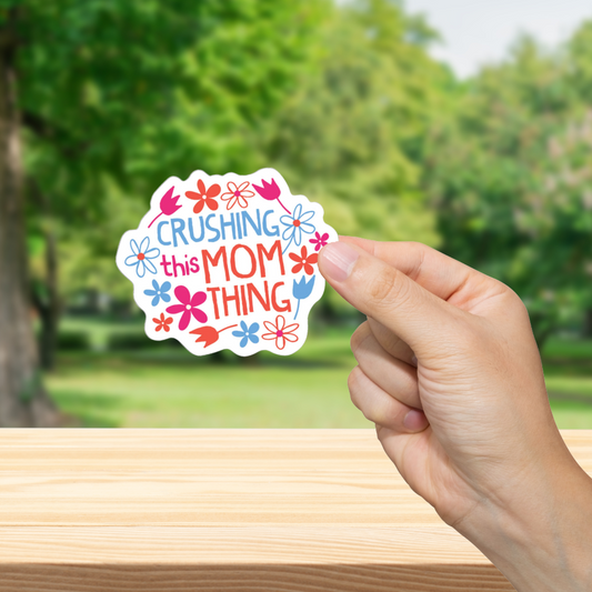 Hand holding a colorful sticker with text "Crushing this Mom Thing." and flowers against a blurred green outdoor background