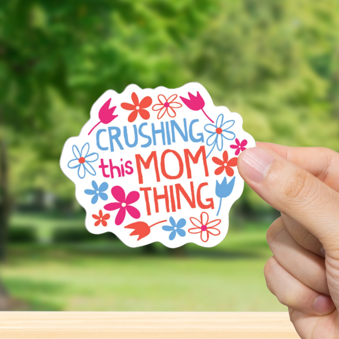 Hand holding a colorful sticker with text "Crushing this Mom Thing." and flowers against a blurred green outdoor background