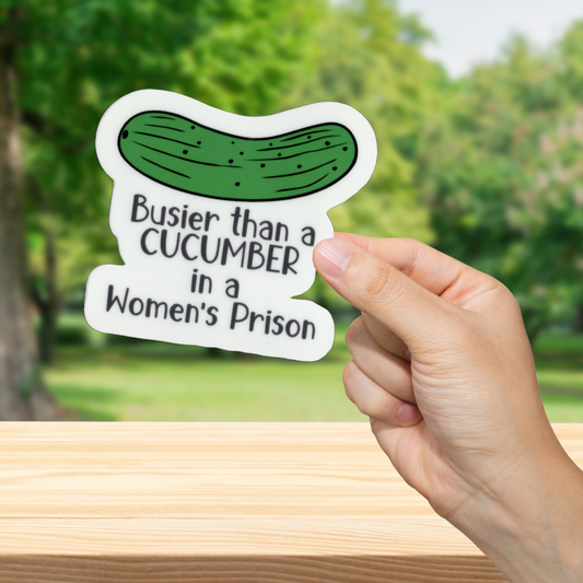 Hand holding a sticker with a pickle graphic and text in front of a blurred green outdoor background