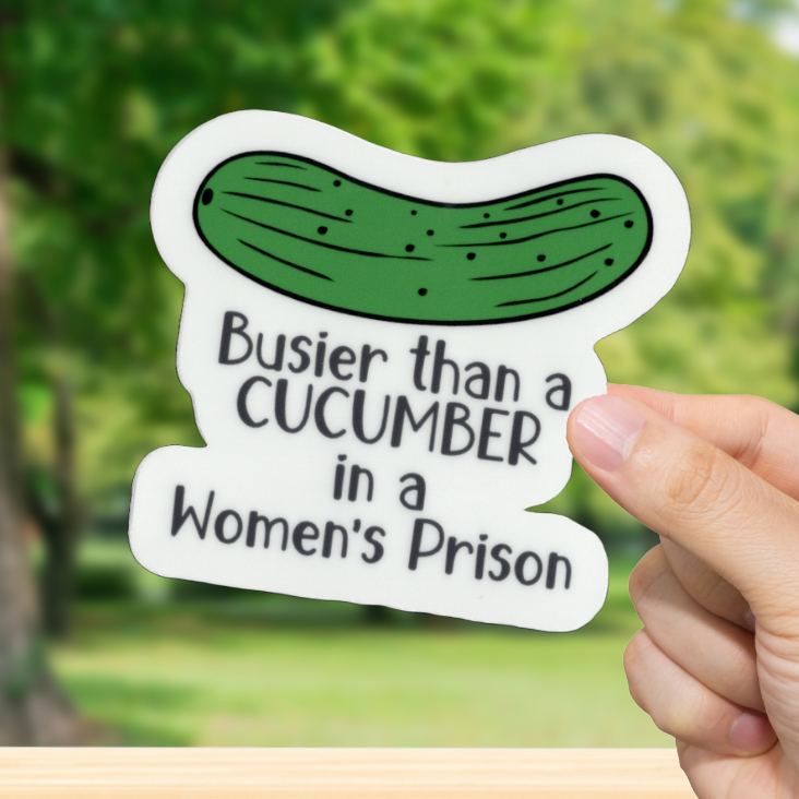 Hand holding a sticker with a pickle graphic and text in front of a blurred green outdoor background