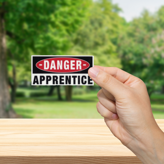 Hand holding a 'Danger: Apprentice' sign with a blurred green outdoor background