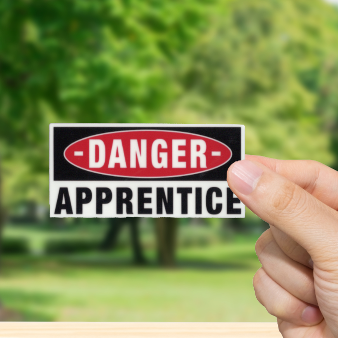 Hand holding a 'Danger: Apprentice' sign with a blurred green outdoor background