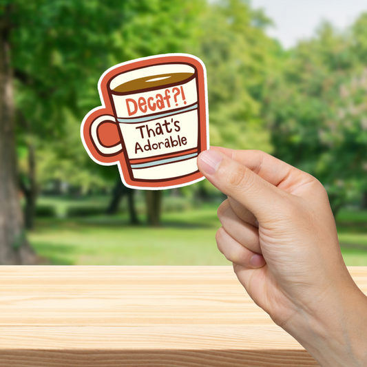Hand holding a sticker with a coffee cup design outdoors. Decaf!? Thats Adorable."