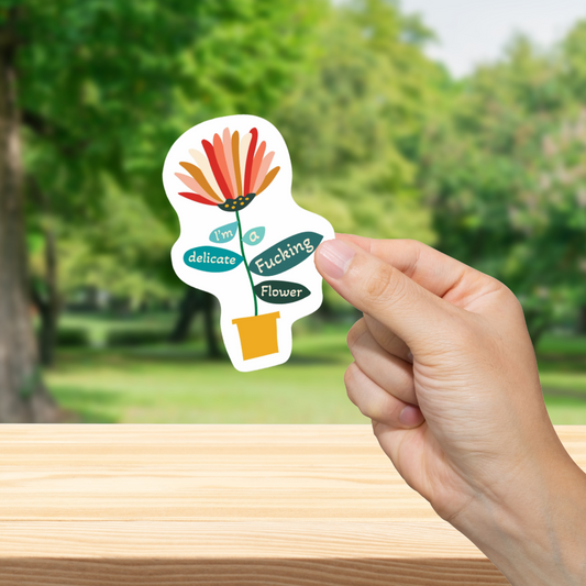Hand holding a sticker with floral design and text "I'm a delicate fucking flower" against a blurred green outdoor background