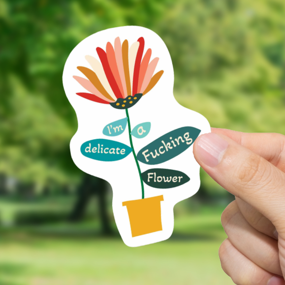 Hand holding a sticker with floral design and text "I'm a delicate fucking flower" against a blurred green outdoor background