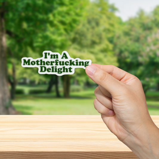 Hand holding a sticker with text 'I'm a mutherfucking delight' in a park setting