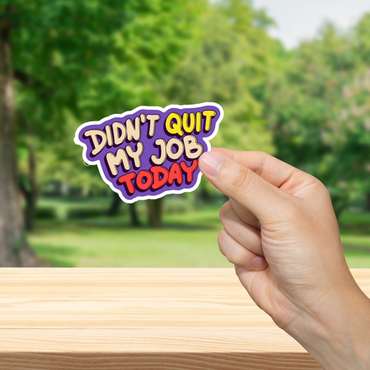 Hand holding a 'Didn't Quit My Job Today' sticker with a blurred green outdoor background