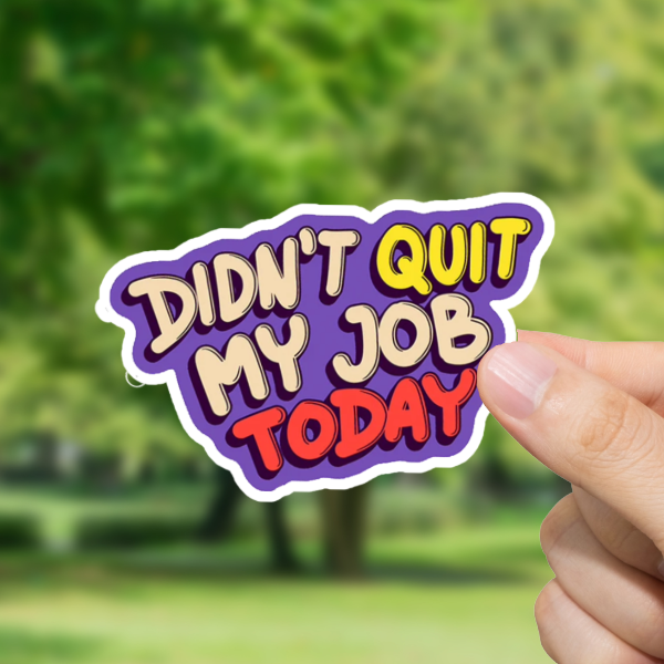 Hand holding a 'Didn't Quit My Job Today' sticker with a blurred green outdoor background