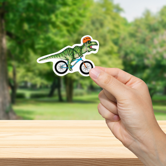 Hand holding a sticker of a dinosaur riding a bicycle with a blurred green outdoor background