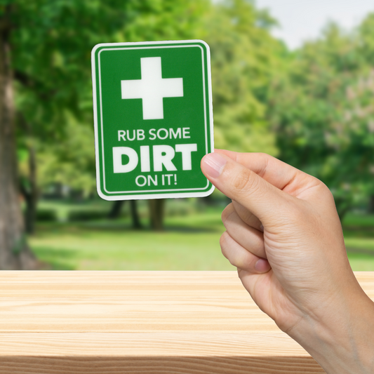 Hand holding a green sign with a cross symbol and text 'Rub some dirt on it' against a blurred outdoor background