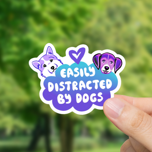 Hand holding a sticker with 'Easily Distracted by Dogs' text against a blurred park background