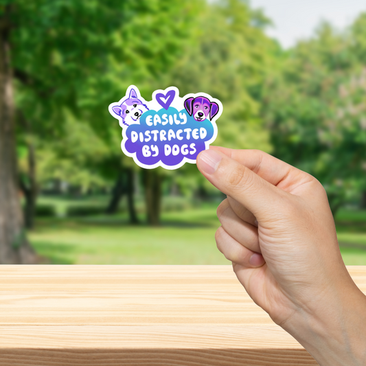 Hand holding a sticker with 'Easily Distracted by Dogs' text against a blurred park background