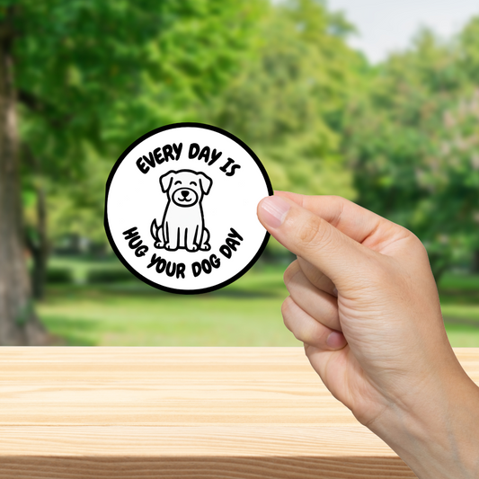 Hand holding a circular sticker with a dog illustration and text 'Every day is hug your dog day' against a blurred green outdoor background
