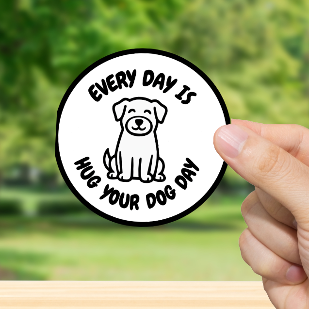 Hand holding a circular sticker with a dog illustration and text 'Every day is hug your dog day' against a blurred green outdoor background