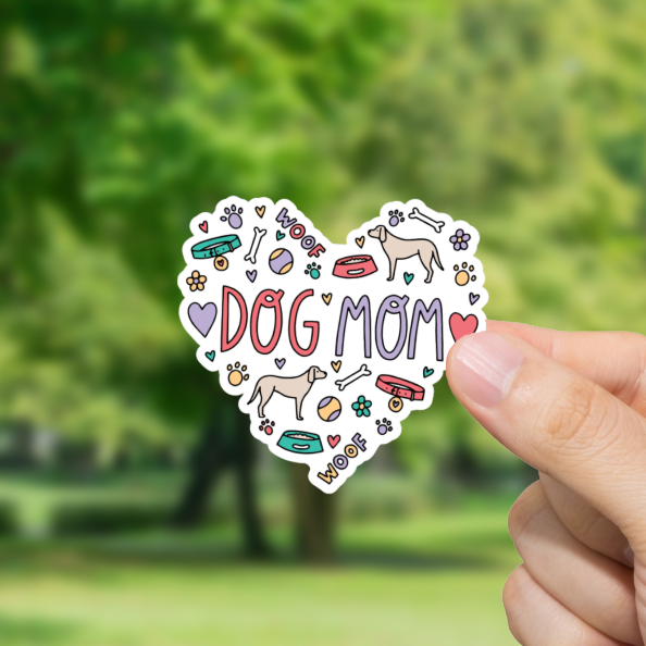 Heart-shaped 'DOG MOM' sticker held by a hand with a blurred green outdoor background