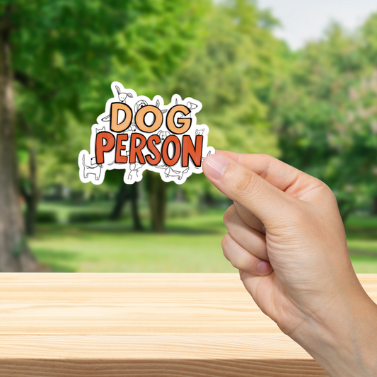Hand holding a 'Dog Person' sticker with a park background