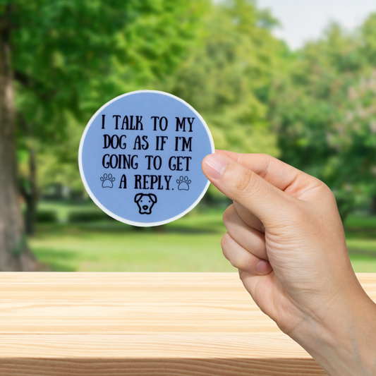 Hand holding a blue sticker with text 'I talk to my dog as if I'm going to get a reply' and a dog icon against a blurred green outdoor background