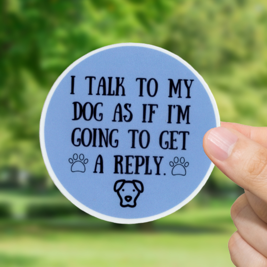 Hand holding a blue sticker with text 'I talk to my dog as if I'm going to get a reply' and a dog icon against a blurred green outdoor background
