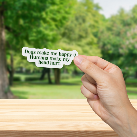 Hand holding a sticker with text "Dogs make me happy, humans make my head hurt' in front of a blurred park background