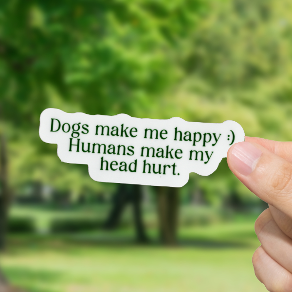 Hand holding a sticker with text "Dogs make me happy, humans make my head hurt' in front of a blurred park background