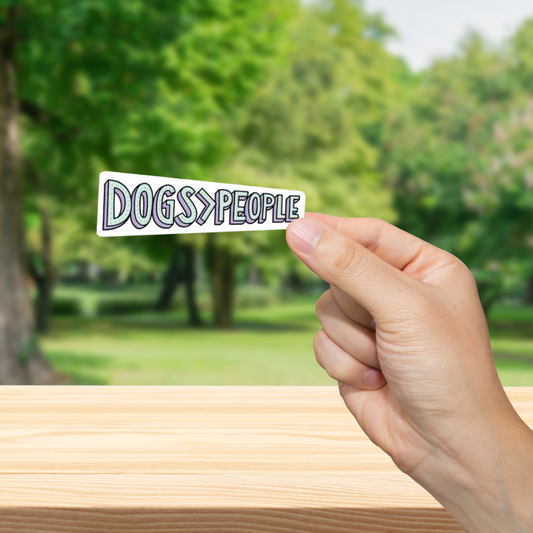 Hand holding a sticker with 'DOGS > PEOPLE' text against a blurred park background