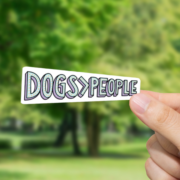 Hand holding a sticker with 'DOGS > PEOPLE' text against a blurred park background