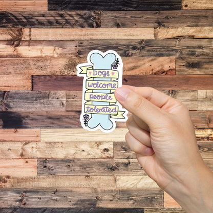 Hand holding a sticker with text 'Dogs welcome, people tolerated' against a wooden background