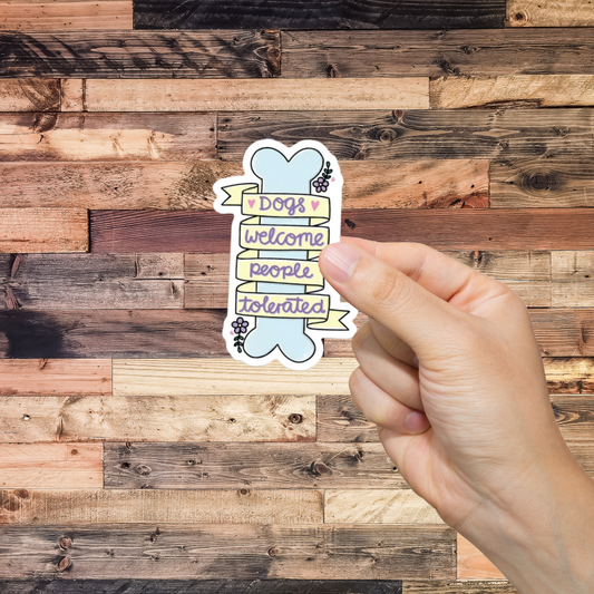 Hand holding a sticker with text 'Dogs welcome, people tolerated' against a wooden background
