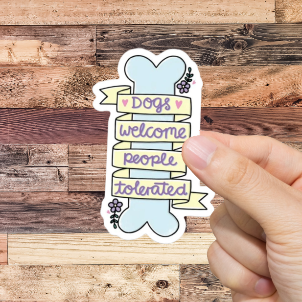 Hand holding a sticker with text 'Dogs welcome, people tolerated' against a wooden background