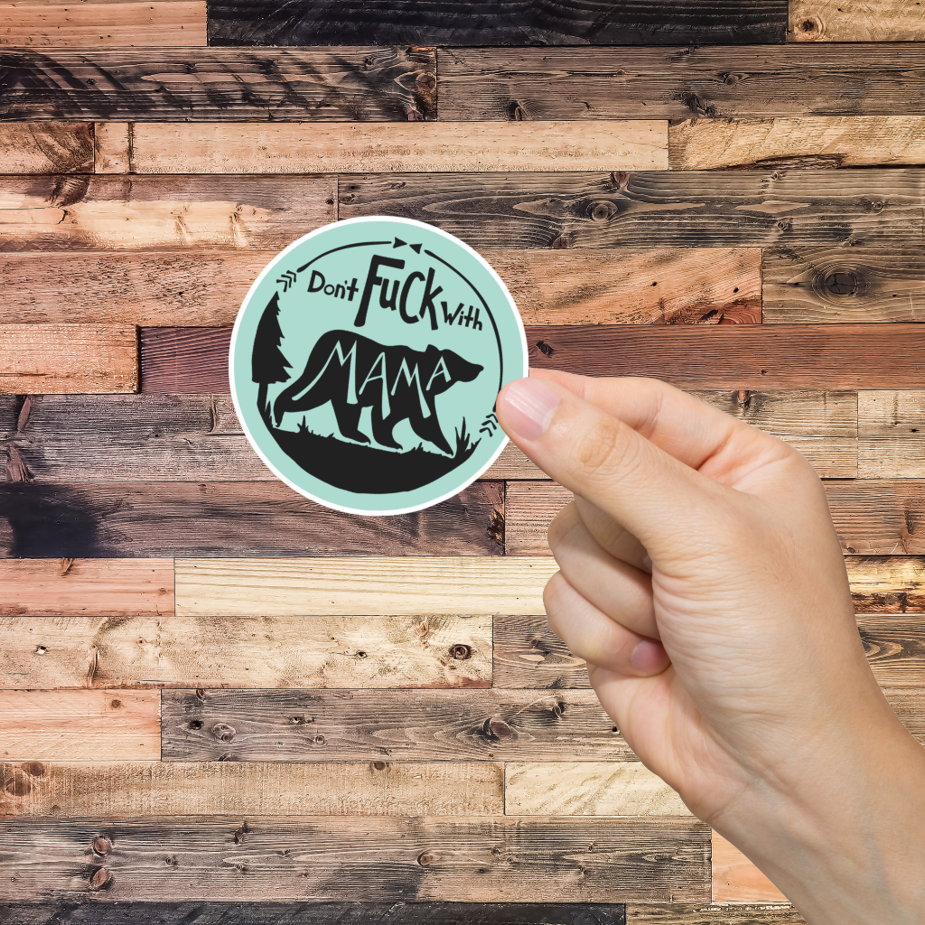 Hand holding a sticker with text 'don't fuck with Mama' and graphic design against a wooden background 