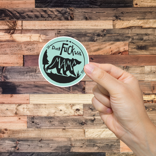 Hand holding a sticker with text 'don't fuck with Mama' and graphic design against a wooden background 