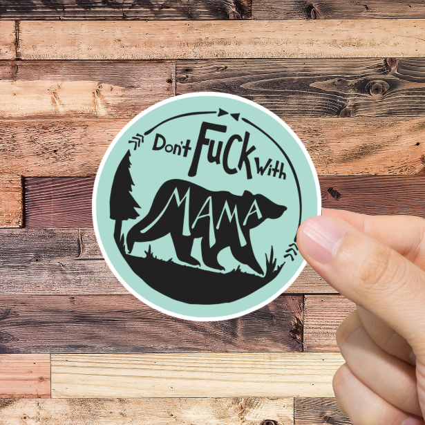 Hand holding a sticker with text 'don't fuck with Mama' and graphic design against a wooden background 