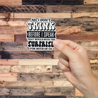 Hand holding a sticker with text 'No, I don't think before I speak. That would ruin the surprise for both of us.' against a wooden background