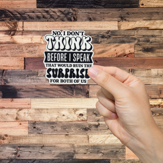 Hand holding a sticker with text 'No, I don't think before I speak. That would ruin the surprise for both of us.' against a wooden background