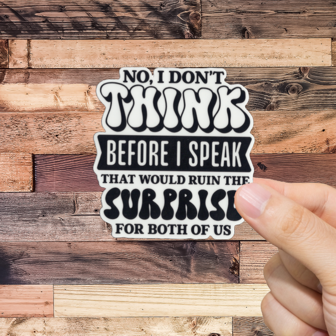 Hand holding a sticker with text 'No, I don't think before I speak. That would ruin the surprise for both of us.' against a wooden background