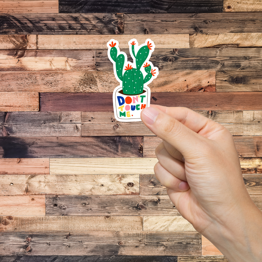Hand holding a sticker with a cactus design and text 'Don't Touch Me' against a wooden background
