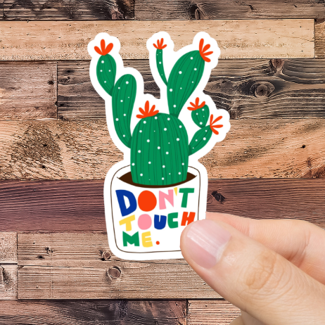 Hand holding a sticker with a cactus design and text 'Don't Touch Me' against a wooden background