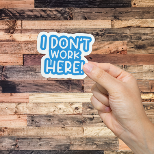Hand holding a sticker with 'I DON'T WORK HERE!' text against a wooden background