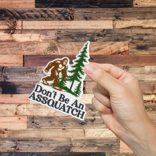 Hand holding a sticker with a Sasquatch design and text 'Don't Be An ASSQUATCH' against a wooden background