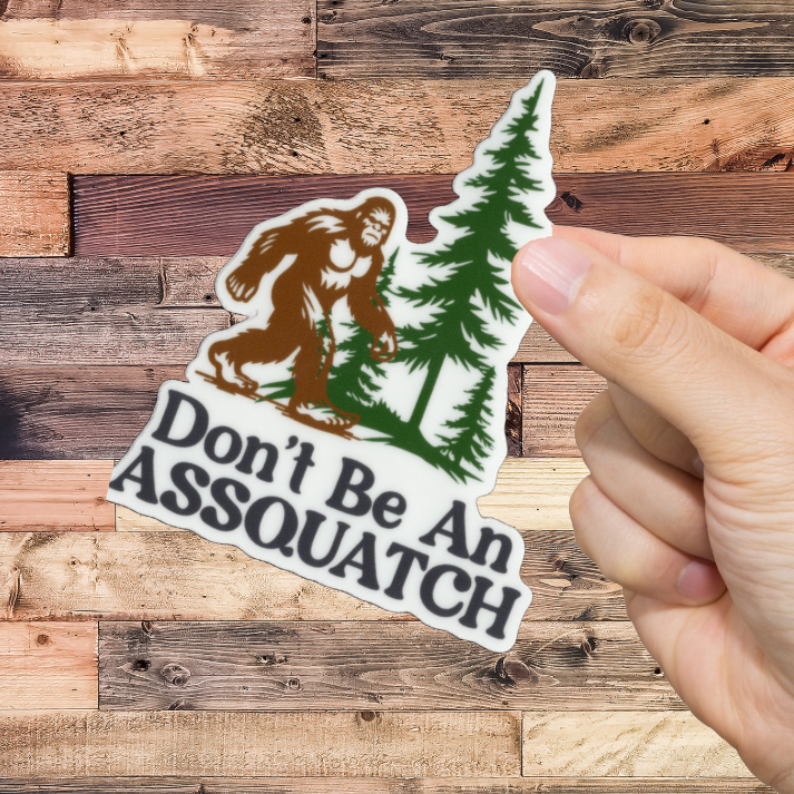 Hand holding a sticker with a Sasquatch design and text 'Don't Be An ASSQUATCH' against a wooden background