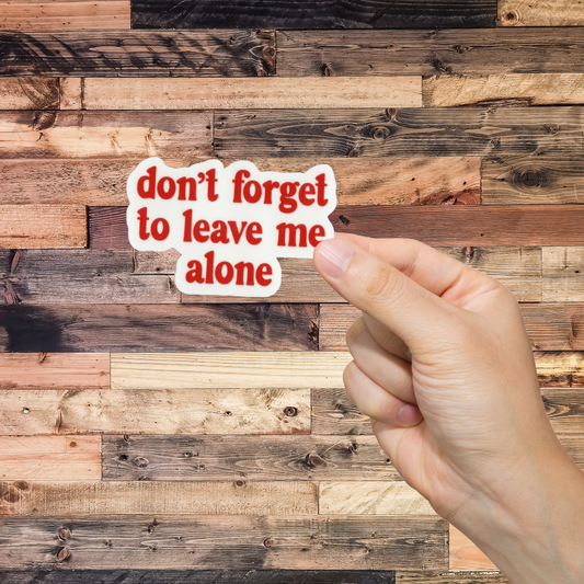 Hand holding a sticker with 'don't forget to leave me alone' text against a wooden background