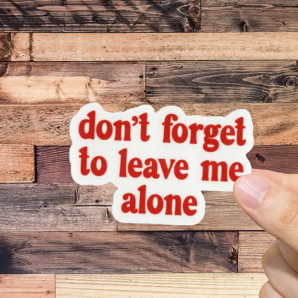 Hand holding a sticker with 'don't forget to leave me alone' text against a wooden background