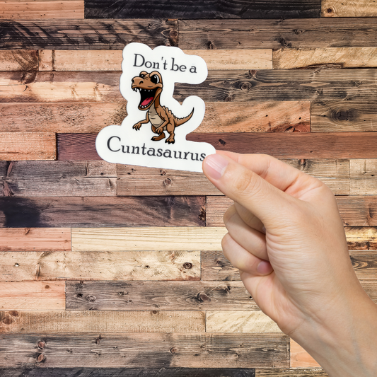 Hand holding a sticker with a dinosaur graphic and text 'Don't be a cuntasaurus' against a wooden background
