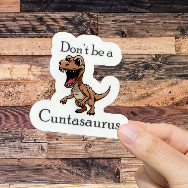 Hand holding a sticker with a dinosaur graphic and text 'Don't be a cuntasaurus' against a wooden background