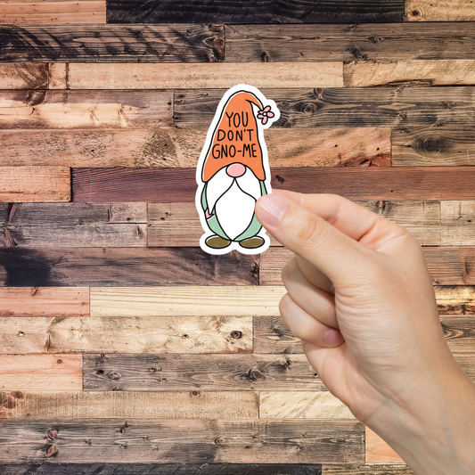 Hand holding a sticker with a gnome and text 'You don't gno-me' against a wooden background