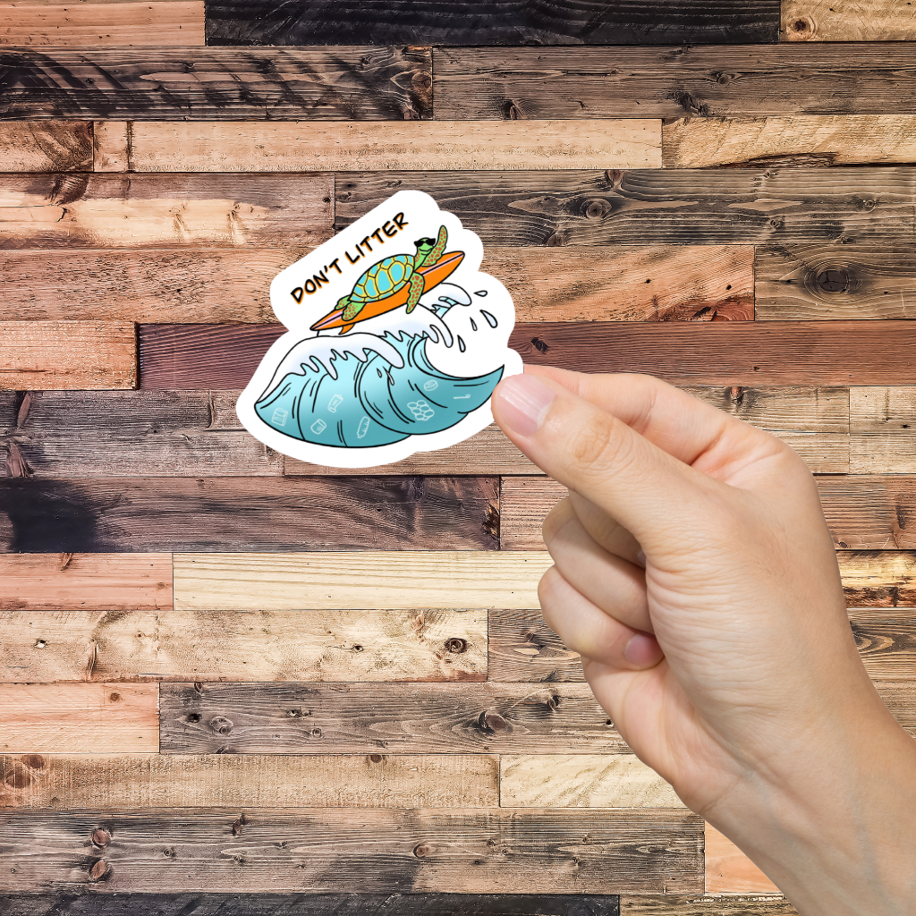Hand holding a sticker with a turtle and wave with 'Don't litter' design against a wooden background
