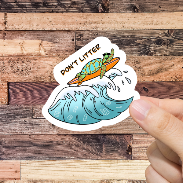 Hand holding a sticker with a turtle and wave with 'Don't litter' design against a wooden background