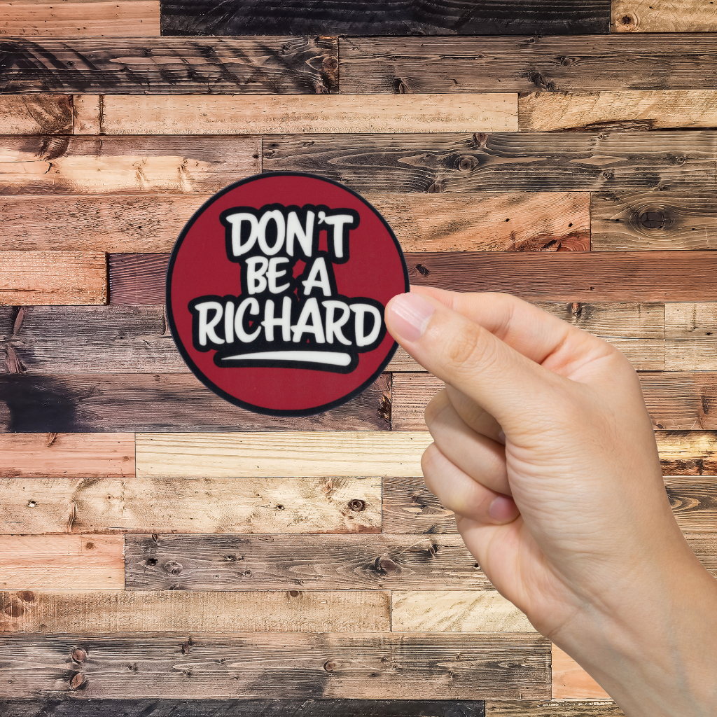Hand holding a round sticker with 'Don't Be A Richard' text against a wooden background